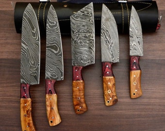 May include: A set of five kitchen knives with Damascus steel blades featuring wavy patterns. Each knife has a two-tone handle made of reddish-purple and light brown wood with brass rivets. The knives are arranged in front of a black leather roll case.