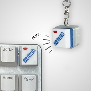 May include: A white keyboard keycap with a red dot and blue Chinese characters that read "Click" on a white keyboard. A white keychain with a blue border and the same Chinese characters is attached to a silver chain. The keychain is labeled "Click" with a red dot.