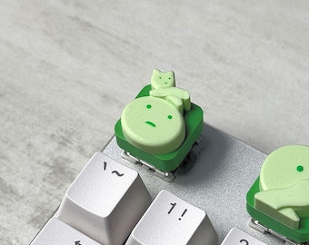 May include: Light green custom keycap with a cartoon-like design of a round face and a small animal on top. The keycap is on a green base, and the keyboard keys are white.