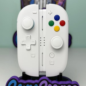 May include: White gaming controller with colored buttons and a directional pad. The controller is on a stand with the text "Game Cosmos" in blue and purple. The stand is black with a purple border.
