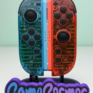 May include: A pair of transparent blue and red game controllers with black buttons, displayed on a stand. The stand is purple and black, with the text "Game Cosmos" in blue and purple. The controllers are designed for a gaming console.