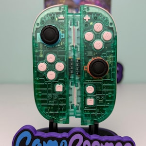 May include: A transparent green video game controller with pink buttons and black joysticks is displayed on a stand. The stand has the words "Game Cosmos" in blue and purple. The controller is designed for a gaming console.