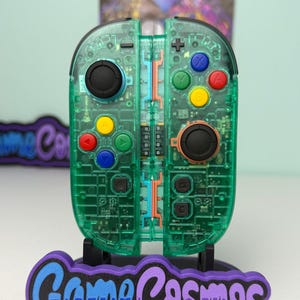 Può includere: Un controller per videogiochi verde trasparente con pulsanti colorati, esposto su un supporto con il testo "Game Cosmos" in blu e viola. Il controller ha joystick neri e un alloggiamento trasparente.