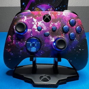 Könnte beinhalten: Ein lila-schwarzer Xbox-Controller mit blauen Knöpfen und einem Galaxie-Design. Der Controller steht auf einem grauen Ständer mit dem Xbox-Logo.