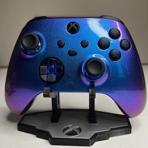 May include: A custom Xbox controller with a gradient blue and purple finish, resting on a matching stand. The controller features black buttons and thumbsticks, with the Xbox logo prominently displayed. The stand is gray and black.