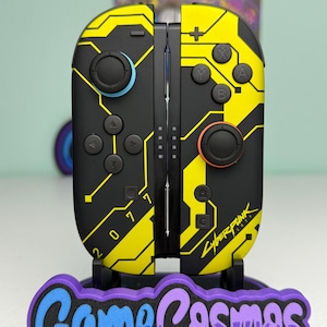 May include: Black and yellow Cyberpunk 2077 themed Nintendo Switch Joy-Cons. The controllers have a circuit board design with the text "2077" and "Cyberpunk". The controllers are on a stand with the text "Game Cosmos".