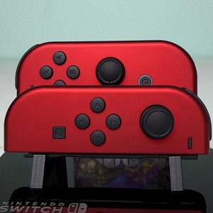 May include: Two red Nintendo Switch Joy-Con controllers on a black stand with the text "NINTENDO SWITCH" printed on the front.