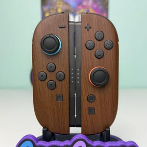 以下が含まれることがあります： 黒いボタンと黒いベースを備えた、木目調の茶色のゲームコントローラーのペア。左側のコントローラーはジョイスティックの周りに青いアクセントがあり、右側のコントローラーはオレンジ色のアクセントがあります。ベースには「Game Cosmos」というテキストが青と紫で表示されています。