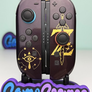 May include: Custom purple Nintendo Switch Joy-Cons with gold Legend of Zelda designs. The controllers have black buttons and joysticks with blue and orange accents. The controllers are on a stand with the text "Game Cosmos".