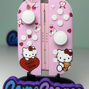 May include: Pink Hello Kitty-themed Nintendo Switch Joy-Con controllers. The controllers feature white buttons, a Hello Kitty graphic, and decorative hearts. The controllers are displayed on a stand with the text "Game Cosmos".