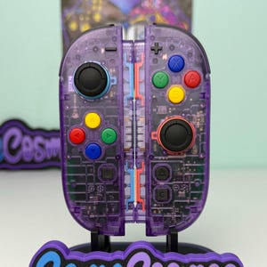 Può includere: Un controller per videogiochi trasparente viola con joystick neri e pulsanti colorati. Il controller è su un supporto con la scritta "Game Cosmos" in blu e bianco. I componenti interni del controller sono visibili.