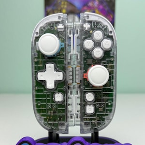 May include: Clear, transparent video game controller with white and red buttons, and a directional pad. The controller is mounted on a stand with the text "Game Cosmos" in blue and purple.