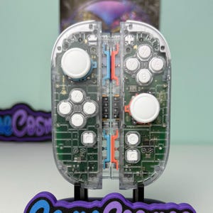 Può includere: Controller di gioco trasparente con componenti interni visibili, pulsanti bianchi e un supporto nero. Il supporto presenta il testo "Game Cosmos" in blu e viola. Progettato per il gaming.