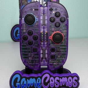 以下が含まれることがあります： 黒いボタンと赤いジョイスティックが付いた、透明な紫色のゲームコントローラー。 コントローラーは、青と紫の「GameCosmos」の文字が入った黒いスタンドに取り付けられています。 コントローラーの内部コンポーネントが表示されます。