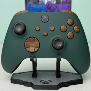 Pode incluir: Um controlador Xbox verde escuro com botões frontais de madeira e um D-pad de madeira. O controlador está em um suporte cinza.