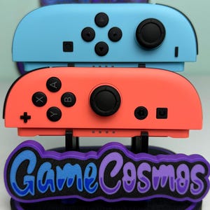 May include: A pair of blue and coral-colored game controllers stacked on a stand with the text "Game Cosmos" in blue and purple. The controllers have black buttons and joysticks.
