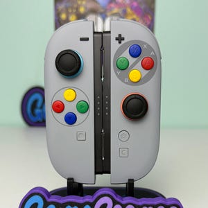 以下が含まれることがあります： 赤、青、黄、緑などのカラフルなボタンが付いたグレーのビデオゲームコントローラー。 コントローラーは、青と紫で「Game Cosmos」というテキストが入ったスタンドに取り付けられています。 スタンドは黒です。