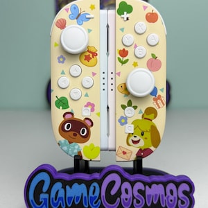 May include: A pair of cream-coloured game controllers featuring a pattern of colourful icons including a blue butterfly, a money bag, a peach, and a gift box. The controllers display cartoon characters Tom Nook and Isabelle on the lower sections.