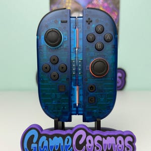 May include: A pair of transparent blue game controllers with black buttons and orange accents, mounted on a stand. The stand features the text "Game Cosmos" in blue and purple against a black base. The controllers are designed for gaming.