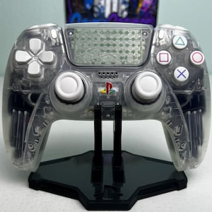 May include: A clear, translucent PlayStation 5 DualSense controller with white buttons and a black PlayStation logo. The controller is on a black stand.