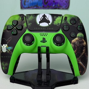 Puede incluir: Un mando de PlayStation 5 verde y negro con un diseño personalizado que presenta al personaje Roronoa Zoro del anime One Piece. El mando tiene un diseño negro y verde con un efecto de humo y una silueta de Zoro en el fondo. El mando está en un soporte negro.