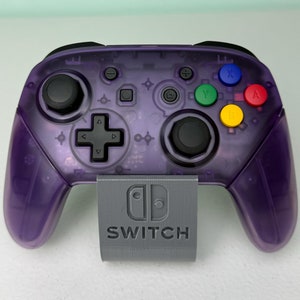 May include: A purple translucent Nintendo Switch controller with a gray stand that says "SWITCH". The controller has a variety of buttons, including A, B, X, Y, and directional buttons.