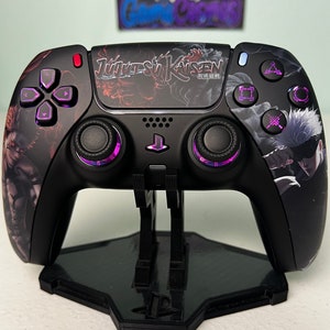 Puede incluir: Un mando de PlayStation 5 DualSense negro con un diseño personalizado de Jujutsu Kaisen. El mando tiene detalles morados en los botones y los gatillos. El mando está en un soporte negro.