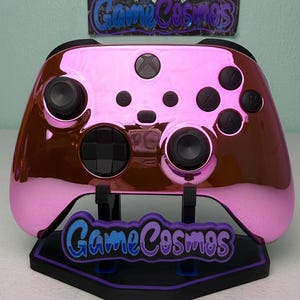 May include: A pink and brown Xbox controller with black buttons and joysticks. The controller is displayed on a black stand with the text "Game Cosmos" in blue and purple. The background is a light teal colour.