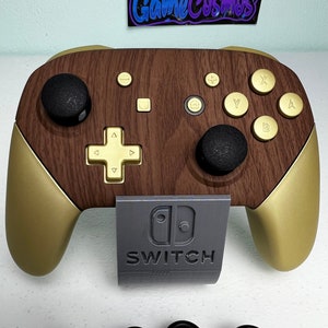 Może przedstawiać: Złoty i brązowy kontroler Nintendo Switch z szarym logo Switch i napisem "SWITCH" nadrukowanym z przodu. Kontroler ma drewnianą fakturę i jest dostarczany z czterema czarnymi gałkami.