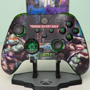 Może przedstawiać: Zielony kontroler Xbox z wzorem Teenage Mutant Ninja Turtles. Kontroler znajduje się na czarnym stojaku z zielonym światłem. Kontroler ma zielono-czarną krzyżówkę i zielone przyciski. Kontroler ma wzór Teenage Mutant Ninja Turtles z napisami "Teenage Mutant Ninja Turtles" i "Turtles" na nim.