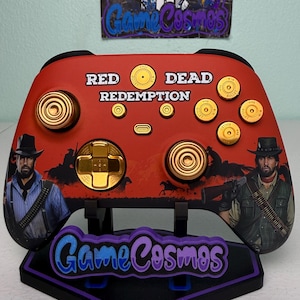 Puede incluir: Mando de videojuegos rojo y dorado personalizado con texto "Red Dead Redemption" y gráficos de personajes. Cuenta con botones dorados y un soporte a juego con el logotipo "Game Cosmos".