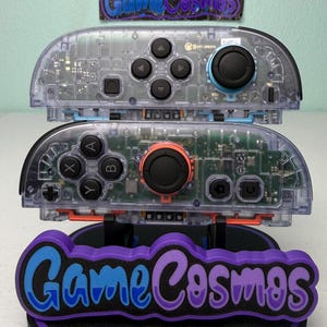 May include: Clear game controllers with visible internal components, black buttons, and colored joysticks. The controllers are stacked on a stand with the text "Game Cosmos" in blue and purple.
