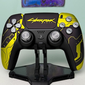 Może przedstawiać: Czarny i żółty kontroler do gier wideo z motywem Cyberpunk 2077. Kontroler ma logo gry i numer 2077 w kolorze żółtym. Kontroler znajduje się na czarnym stojaku.