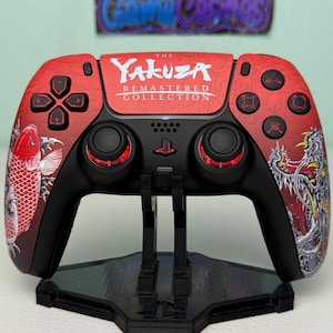 May include: A black PlayStation 5 DualSense controller with a custom design featuring a red and white koi fish and a dragon on a red background. The controller has the text "The Yakuza Remastered Collection" on the front.