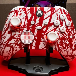 Puede incluir: Un mando Xbox blanco y rojo con un diseño de pintura salpicada. El mando tiene un pad direccional blanco, botones blancos y gatillos blancos. El mando está en un soporte negro.
