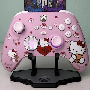 May include: A pink Xbox controller featuring a Hello Kitty design. The controller has a pink and white Hello Kitty character on the front, with the words "Hello Kitty" printed on the side. The controller is displayed on a black stand.