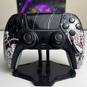 May include: A black and white custom designed PlayStation 5 DualSense controller with a stand. The controller has a black and white design with a red and white graphic on the back. The stand is black and has a PlayStation logo on the front.