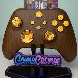 Puede incluir: Un mando Xbox de madera marrón con botones dorados y un pad direccional dorado. El mando está en un soporte negro con las palabras "GameCosmos" impresas en él.
