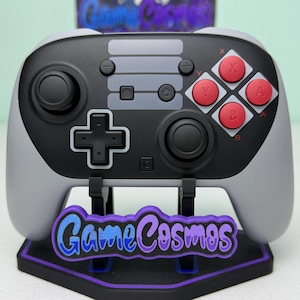 Può includere: Un controller per videogiochi grigio e nero con pulsanti rossi, appoggiato su un supporto con la scritta "Game Cosmos" in viola e blu. Il controller ha un design classico con un D-pad, pulsanti e levette analogiche.