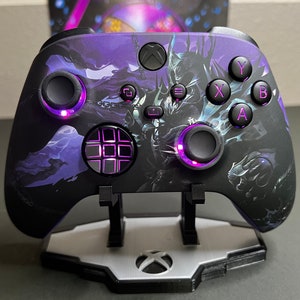 Könnte beinhalten: Ein schwarzer Xbox-Controller mit einem lila und schwarzen Design, das einen Fantasy-Charakter zeigt. Der Controller hat ein lila Licht um die Tasten und das Steuerkreuz.