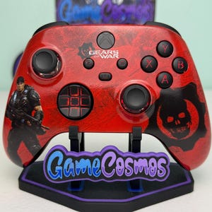 Może przedstawiać: Czerwono-czarny kontroler Xbox Gears of War z teksturowanym wzorem. Zawiera grafikę postaci, logo czaszki i logo gry. Kontroler jest wyświetlany na stojaku z tekstem "Game Cosmos".