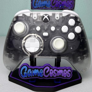 May include: A transparent Xbox controller with white buttons and a black Xbox logo. The controller sits on a black stand with the text "Game Cosmos" in blue and purple. The controller's internal components are visible.
