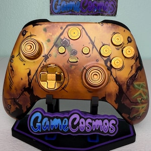 Peut inclure: Manette de jeu vidéo personnalisée avec une finition dorée et marron vieillie. Elle présente des boutons et des accents dorés, ainsi qu'un support avec le texte "Game Cosmos". Le design de la manette est artistique et unique.