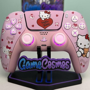 May include: A light pink wireless video game controller featuring Hello Kitty graphics, including a red heart with the text Hello Kitty, cherries, and envelopes. The controller has white buttons, illuminated analog sticks, and rests on a black stand.