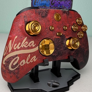 以下が含まれることがあります： ゴールドのボタンとヌカ・コーラのロゴが付いた赤と黒のXboxコントローラー。コントローラーはスタンドの上に置かれています。