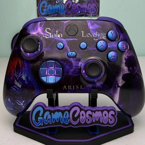Puede incluir: Mando de Xbox personalizado con un diseño de Solo Leveling. El mando presenta una combinación de colores púrpura y negro con un patrón similar a una nebulosa y la palabra "ARISE". Se muestra en un soporte a juego con el logotipo de "Game Cosmos".