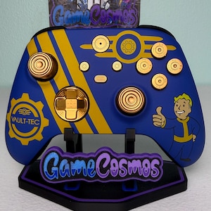 May include: A custom blue and yellow video game controller with gold accents and a Vault-Tec logo. The controller features a thumbs-up character and the text "Game Cosmos" on the stand.