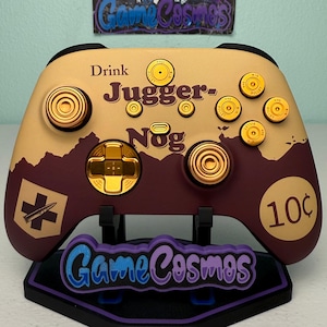 May include: A custom video game controller featuring a tan and maroon color scheme with gold-colored buttons and joysticks. The controller displays the text Drink Jugger-Nog, a bullet icon, and a 10 cent symbol. The GameCosmos logo is visible.