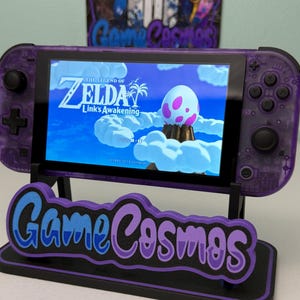 Könnte beinhalten: Eine lila Nintendo Switch-Konsole, die "The Legend of Zelda: Link's Awakening" auf dem Bildschirm anzeigt, ruht auf einem schwarzen Ständer mit dem "Game Cosmos"-Logo in Blau und Lila. Die Konsole hat ein transparentes lila Gehäuse.
