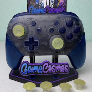 May include: A translucent blue and gray wireless video game controller with yellow thumbsticks, mounted on a black stand with the text GameCosmos. Four additional yellow thumbstick caps are placed in front of the stand.